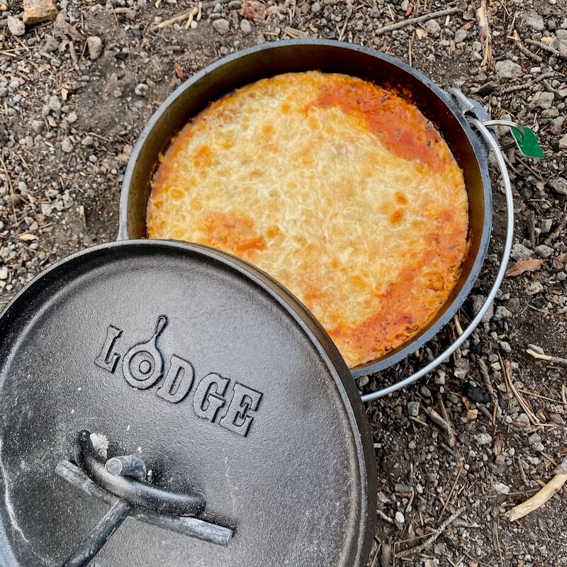 A camp Dutch oven filled with lasagna, freshly cooked over hot coals, showcasing the versatility of outdoor cast iron cookware.