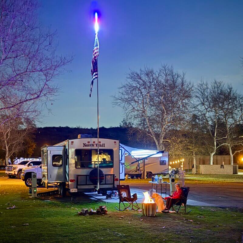 RV enthusiast relaxing by a campfire at a scenic campsite, with an American flag flying high on a truck flag mount attached to a pickup towing an RV, under a starry night sky.