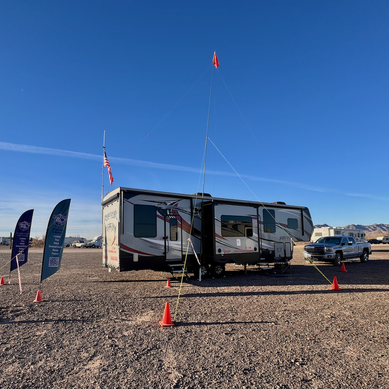 RV parked in a desert campsite with an MFJ mast mounted on a tire mount, American flag, and event banners under a clear blue sky.